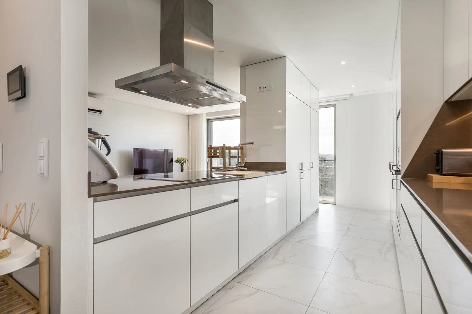 Cozinha moderna com balcões brancos, armários de armazenamento, cooktop de indução, exaustor, janela grande com vista urbana ao fundo, piso de mármore, e utensílios de cozinha minimalistas.