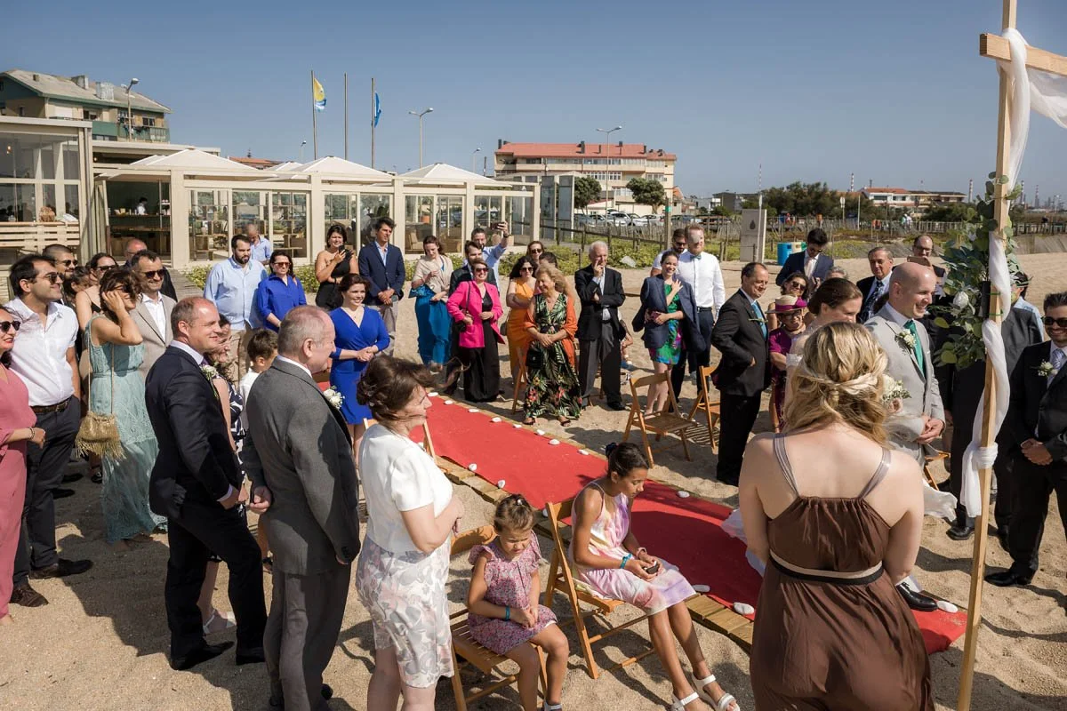 Casal de casamento caminando na direção de um altar na praia, com convidados ao seu redor assistindo ao casamento ao ar livre.