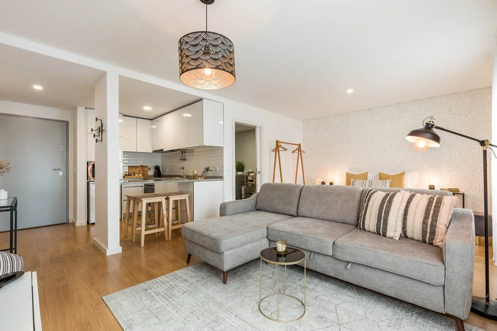 Sala de estar moderna com sofá cinza, mesa de centro pequena, iluminação de teto decorativa, e espaço de jantar ao fundo com cadeiras de madeira e uma cozinha branca ao lado.