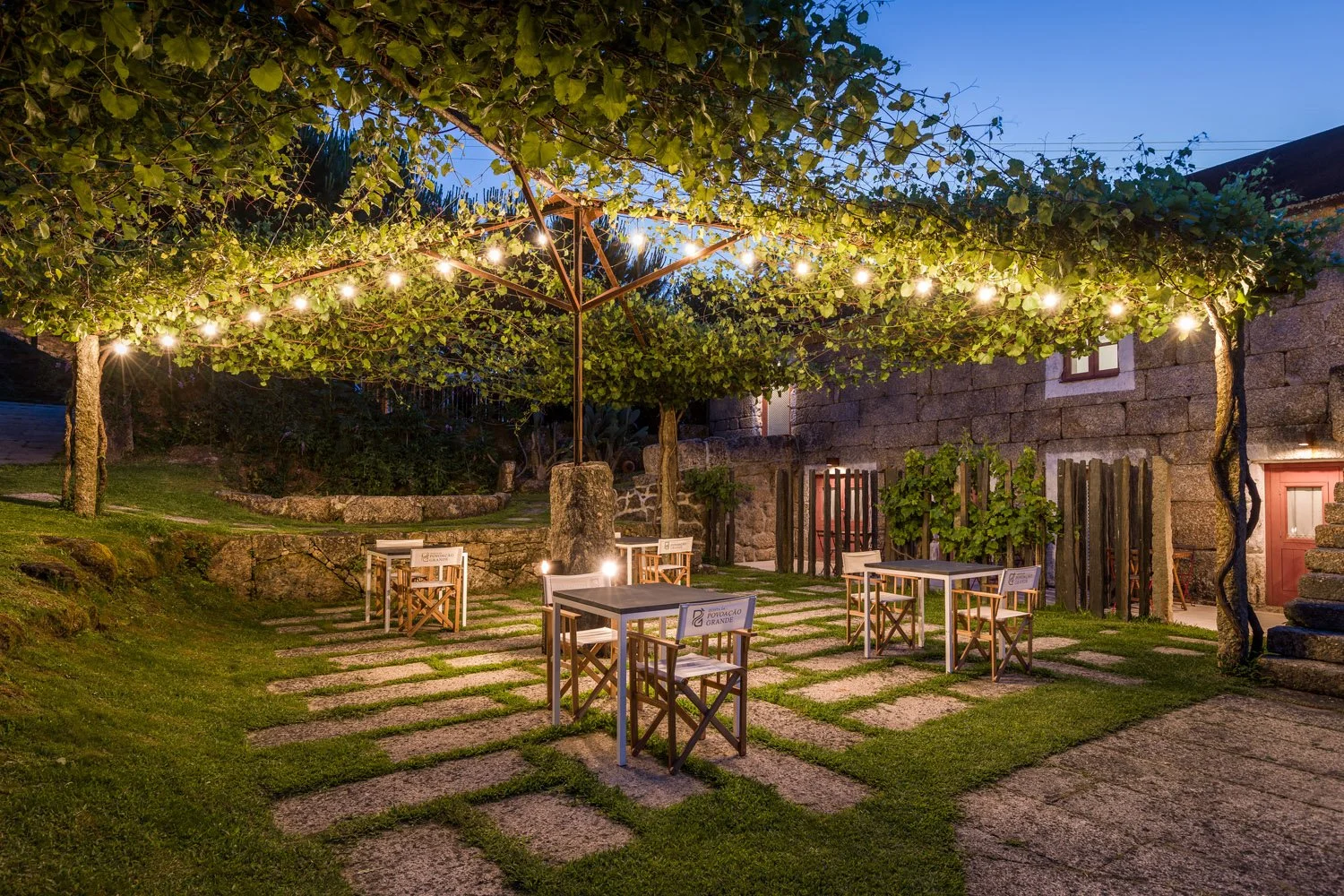 Ilha ao ar livre com mesas e cadeiras, teto de vegetação com luzes penduradas, ambiente ao entardecer ou noite, cercado por vegetação e paredes de pedra.