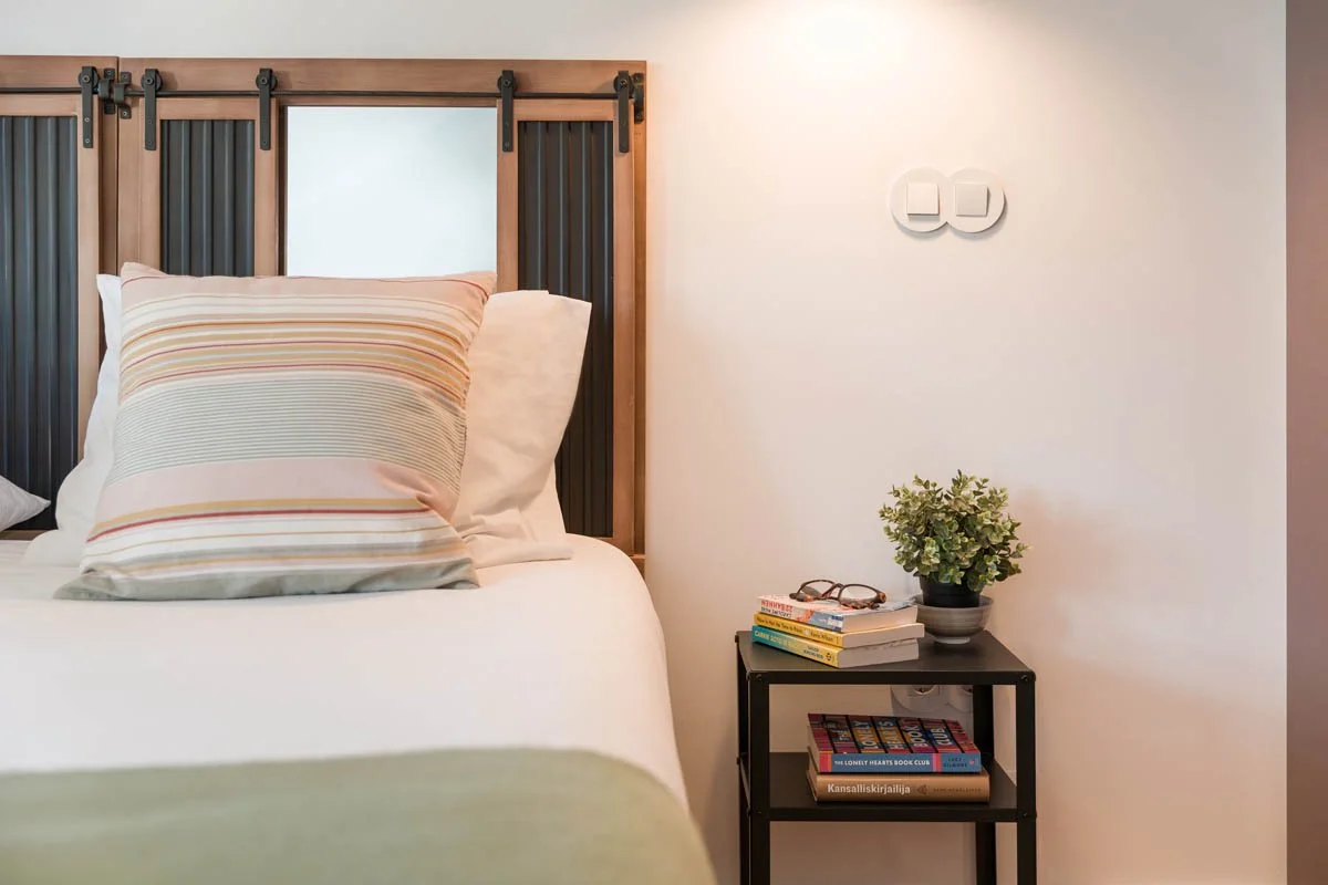 Cama com cabeceira de madeira e ripas pretas, travesseiro com capa de cama listrada em cores suaves, ao lado uma mesa preta com livros, óculos, uma planta e interruptores na parede branca.