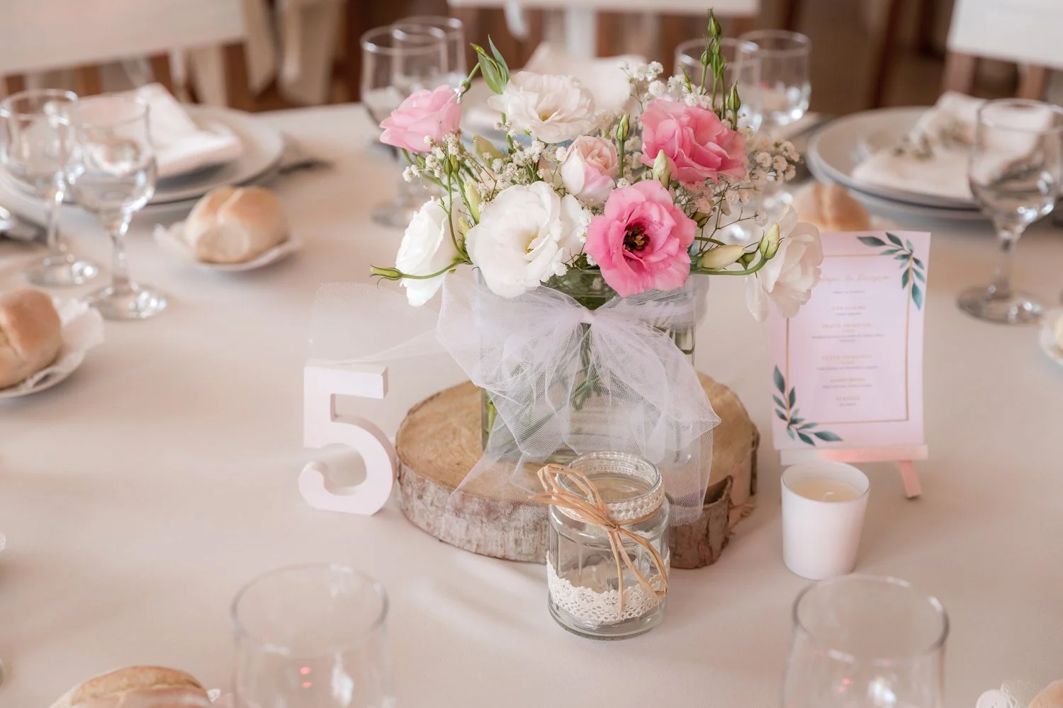 Mesa decorada com vaso de flores em tons de rosa e branco, com um número 5 de decoração, velas, pão, cardápio e taças de vidro, em um ambiente de celebração ou casamento.