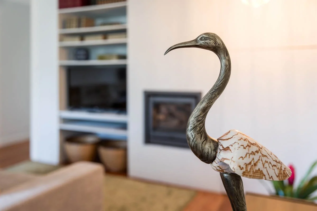 Estátua decorativa de um pássaro semelhante a uma garça com bico longo, colocada em um móvel na sala de estar com lareira ao fundo.