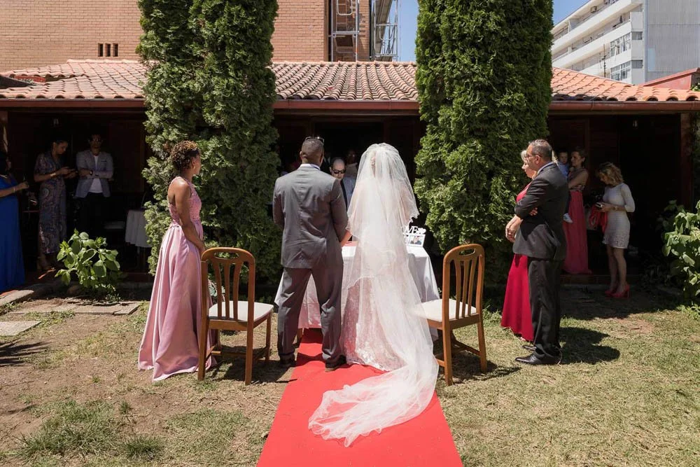 Casamento ao ar livre com noivos, convidados e testemunhas, em um jardim cercado por árvores e uma casa com telhado de telhas. A noiva usa vestido de casamento com véu e o noivo usa terno cinza.
