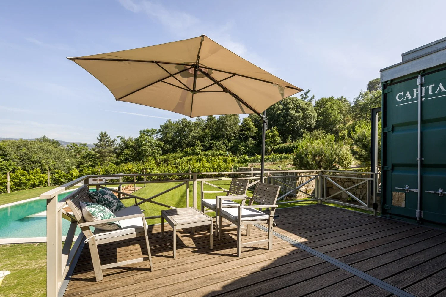 Varanda com mesa, cadeiras, almofadas e guarda-sol, vista para o jardim com piscina e área verde ao fundo.