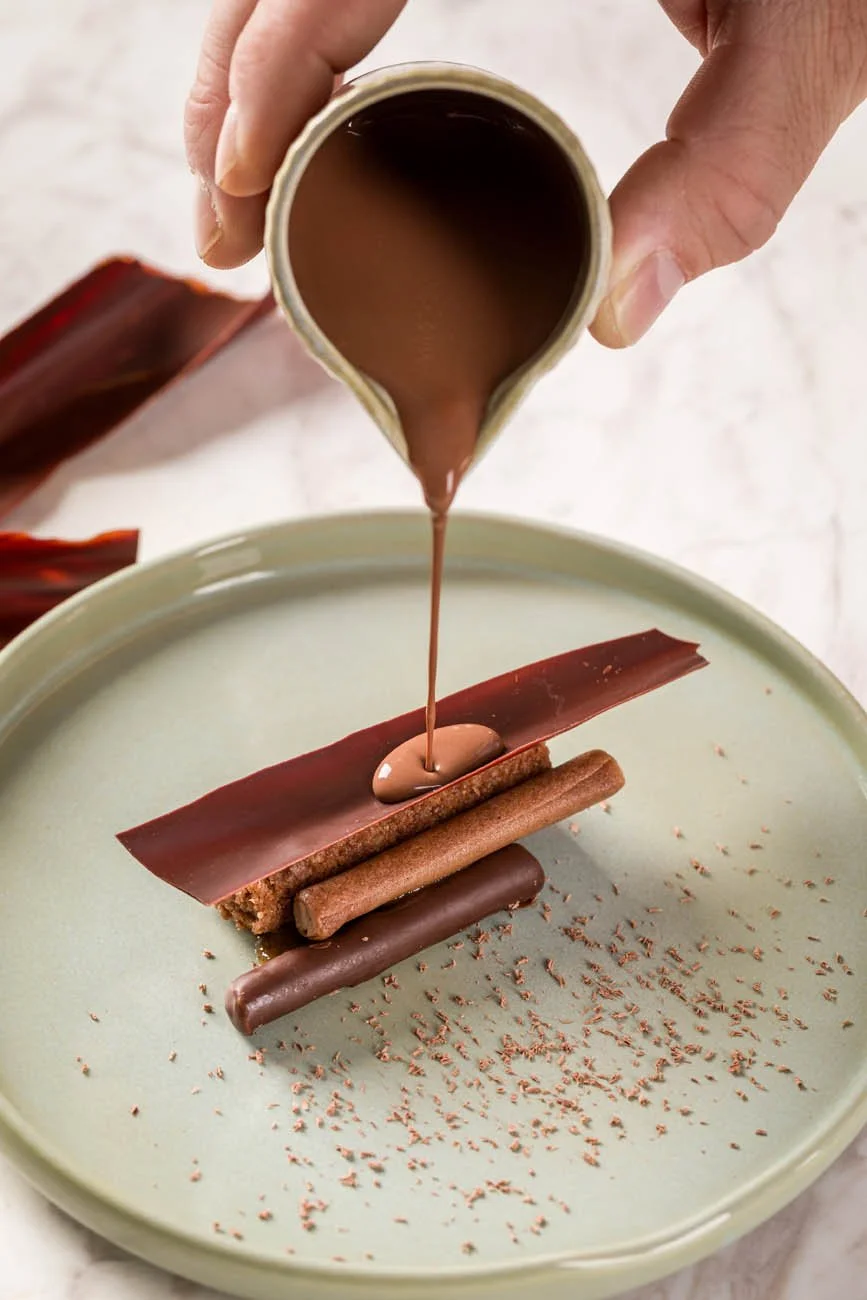 Alguém despejando calda de chocolate sobre uma sobremesa decorada com diversos chocolates e ingredientes de confeitaria em um prato de porcelana.
