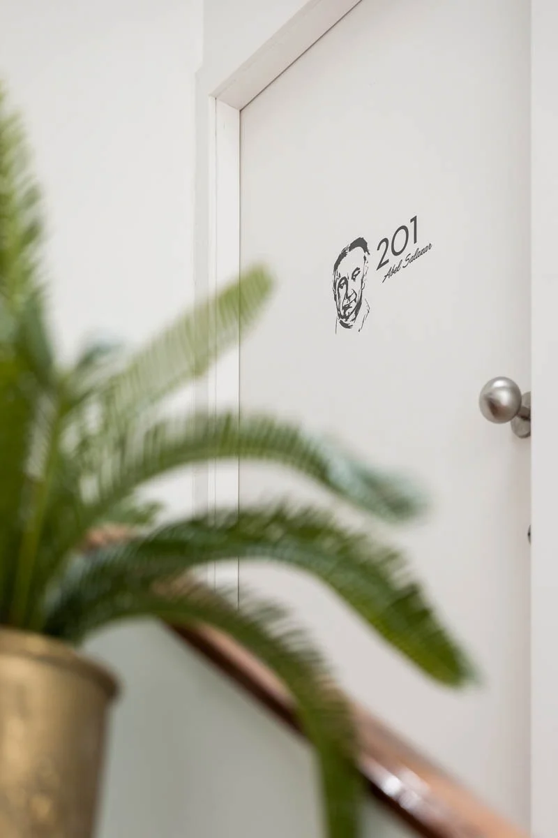 Porta branca com etiqueta 201, desenho de um rosto e assinatura ao lado. Planta de folhas verdes ao fundo.