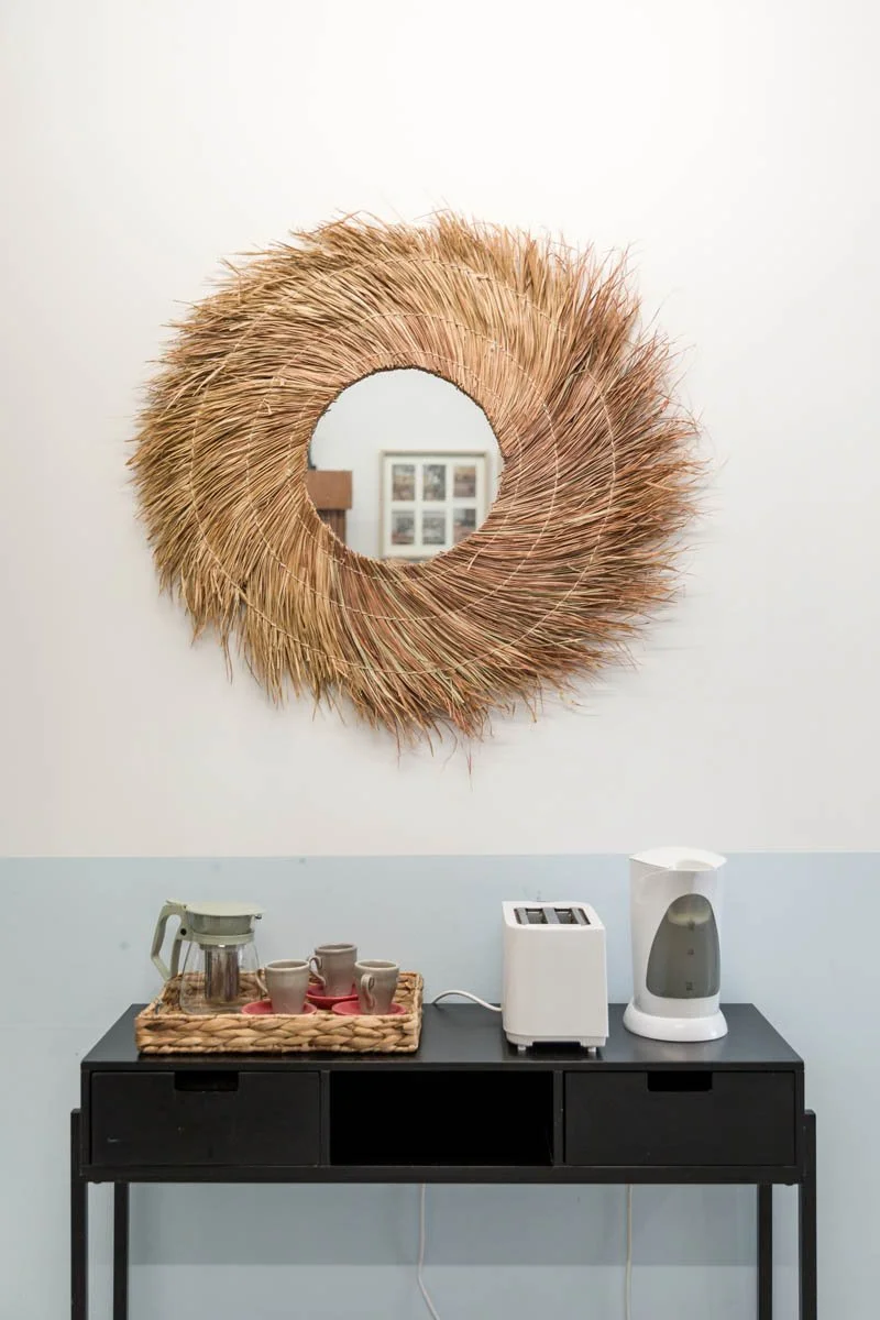 Espelho decorativo redondo feito de palha e fibra natural, colocado na parede branca acima de um móvel preto com utensílios e máquina de café.