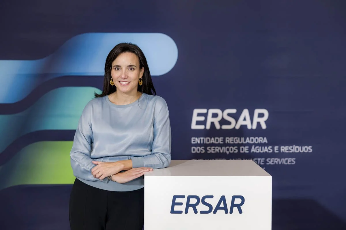 Mulher sorridente apoiada sobre uma mesa branca com o logotipo 'ERSAR' em fundo azul escuro com texto em português e inglês