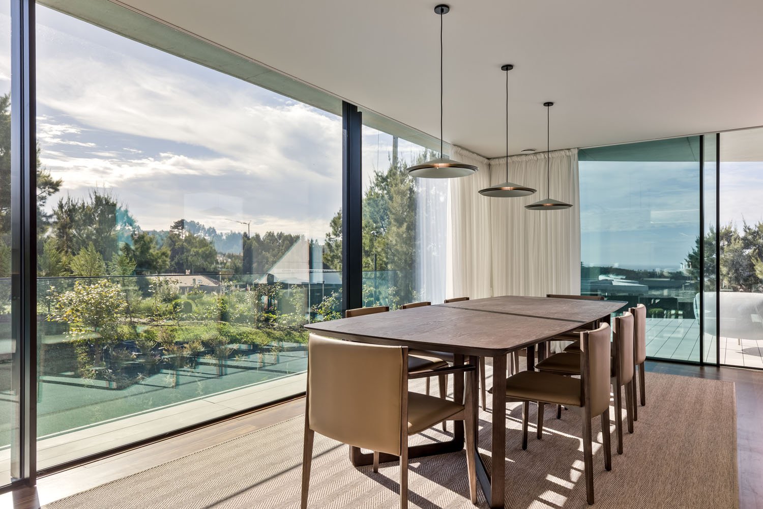 Sala de jantar moderna com mesa de madeira e oito cadeiras em ambiente com janelas de vidro e vista para o jardim externo e céu nublado.