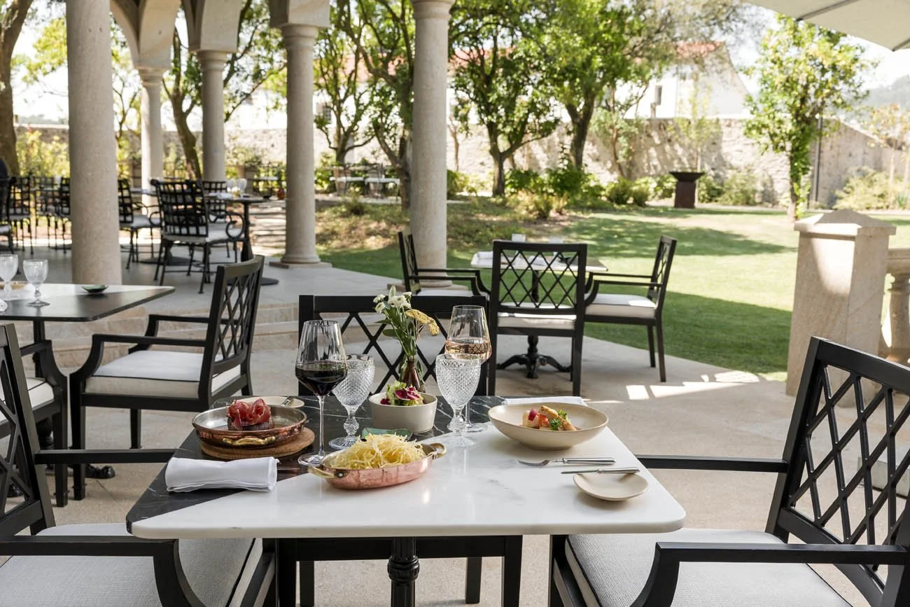 Mesa de jantar ao ar livre com pratos de comida, taças de vinho, água e flores em um ambiente externo com colunas e árvores ao fundo.