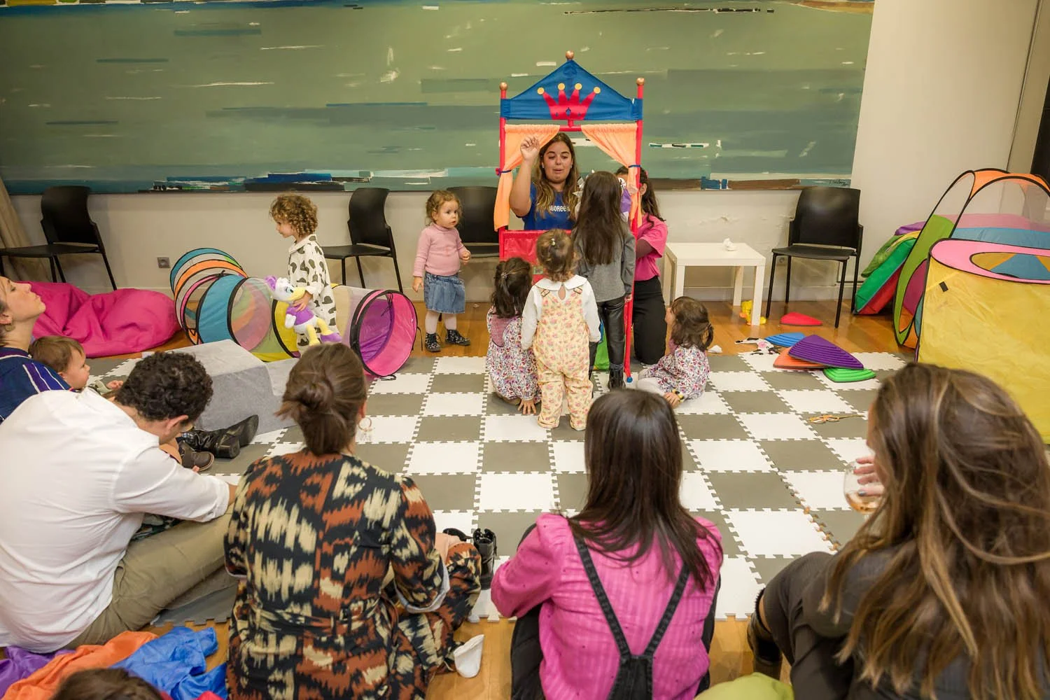 Crianças e adultos assistindo uma peça de teatro infantil com um palhaço em um espaço com tapete de borracha e brinquedos coloridos ao redor.