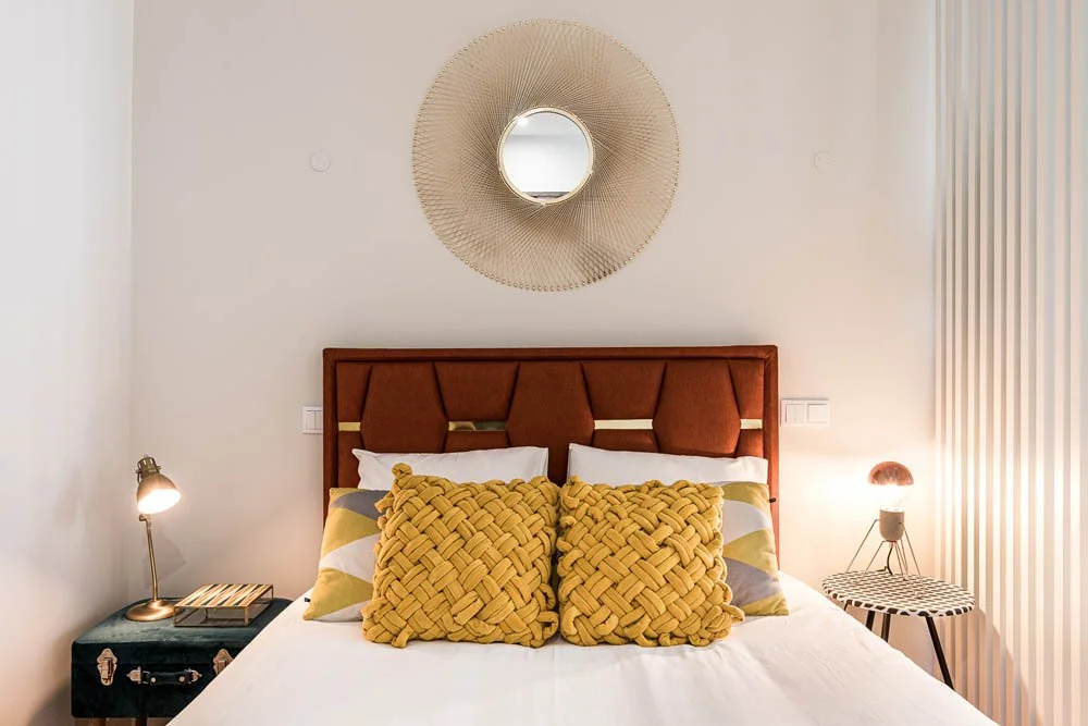 Quarto com cama, almofadas amarelas ou beges, espelho decorativo sobre a cabeceira, luminária de um lado, abajur no outro lado, parede branca e detalhes de decoração floral ou de padrão nas paredes.
