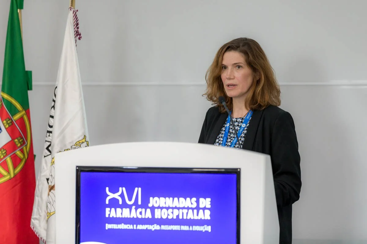 Mulher falando em um evento de farmácia hospitalar, com bandeiras ao fundo e uma tela digital na frente com o texto "XVI JORNADAS DE FARMÁCIA HOSPITALAR".