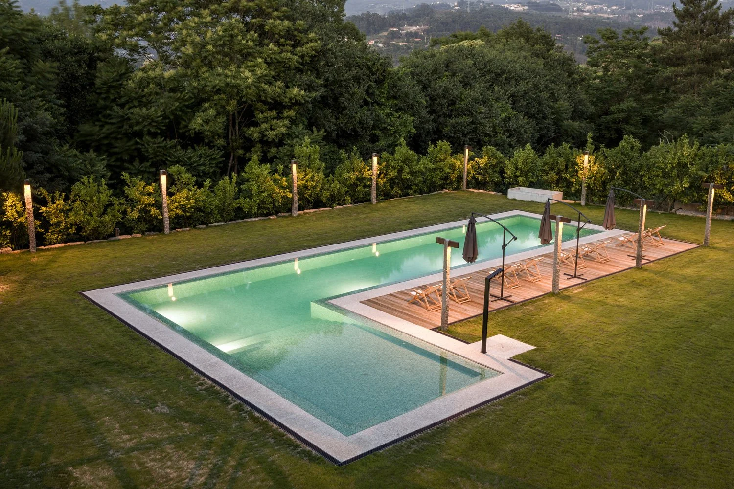 Piscina ao ar livre com deck de madeira, cadeiras reclináveis, guarda-sóis, iluminação ao redor, cercada por árvores e vegetação verde, ao entardecer.