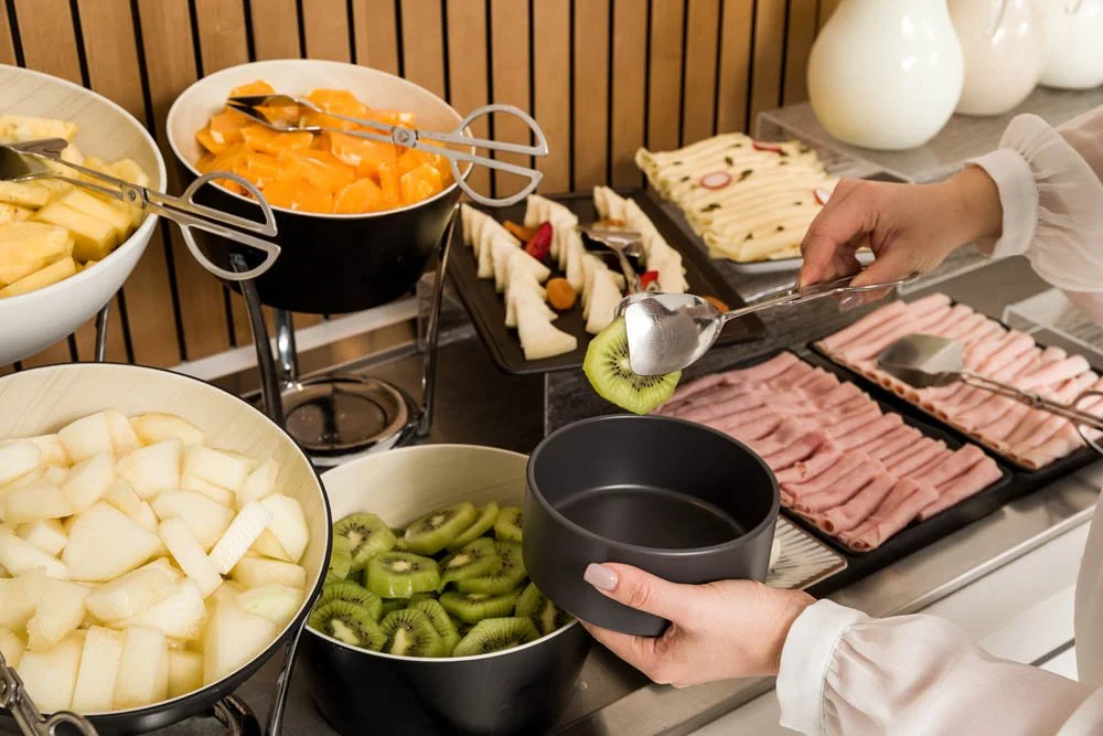 Buffet com frutas variadas, incluindo melão, kiwi, melancia e mamão, além de fatias de presunto, queijo e pão.