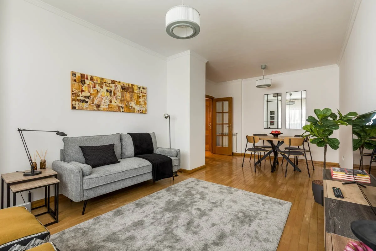 Sala de estar com sofá cinza, mesa de centro, tapete, parede branca, obras de arte na parede e plantas verdes