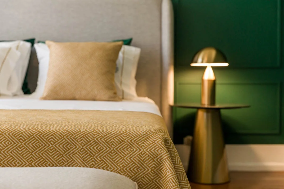 Cama com colcha amarela e travesseiros, ao lado de uma luminária dourada em mesa de cabeceira ao lado de uma parede verde