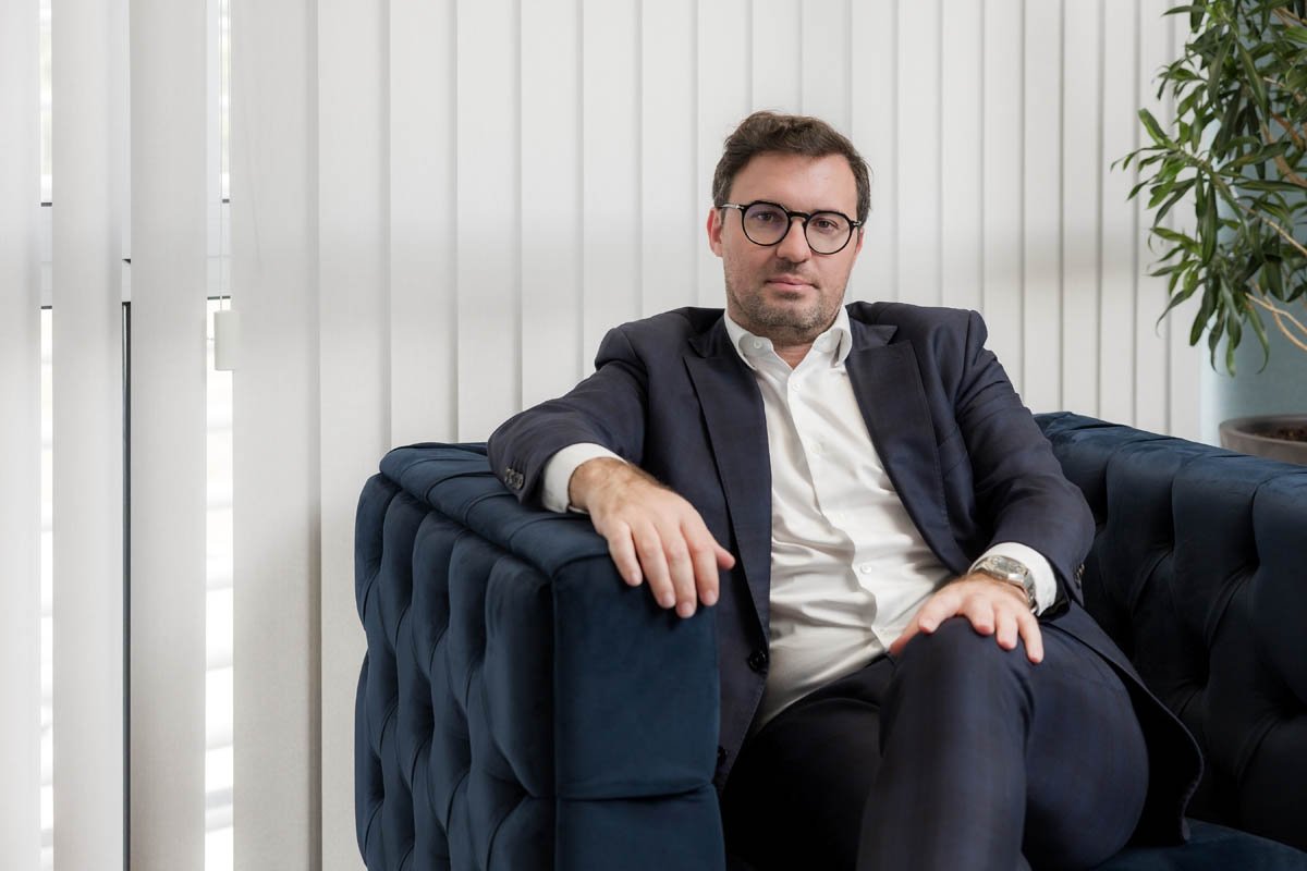Homem de terno escuro sentado em um sofá azul, com janelas de persianas brancas ao fundo e planta ao lado.