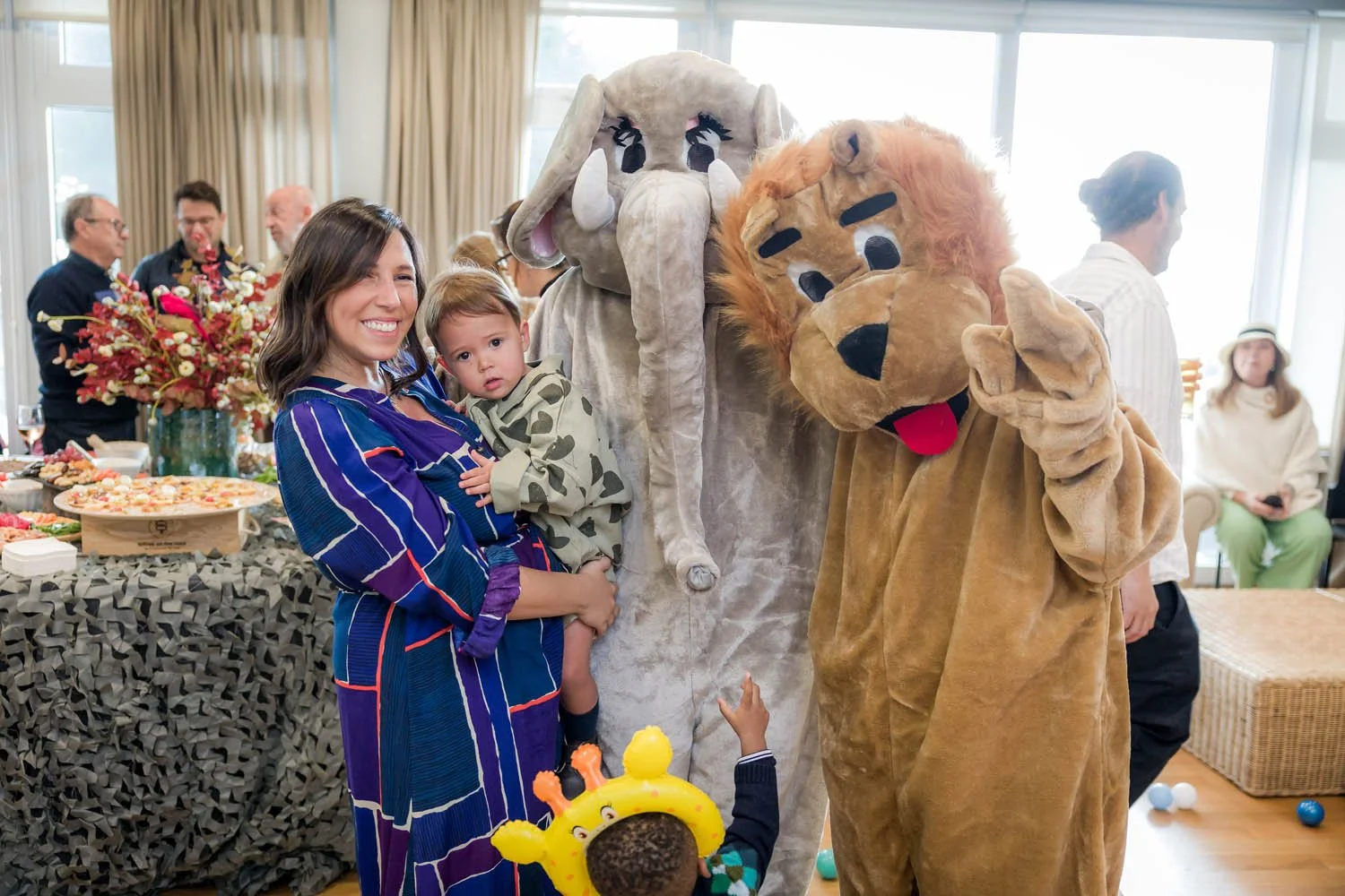 Pessoas em uma festa com crianças e dois personagens fantasiados de animais, um elefante e um leão, em um ambiente bem iluminado com uma mesa de comidas e pessoas ao fundo.