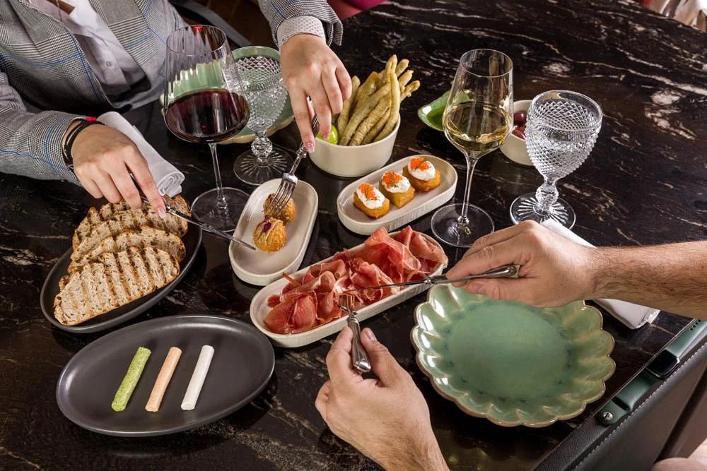Mesa de jantar com tapas, vinhos e água, com pessoas servindo-se.