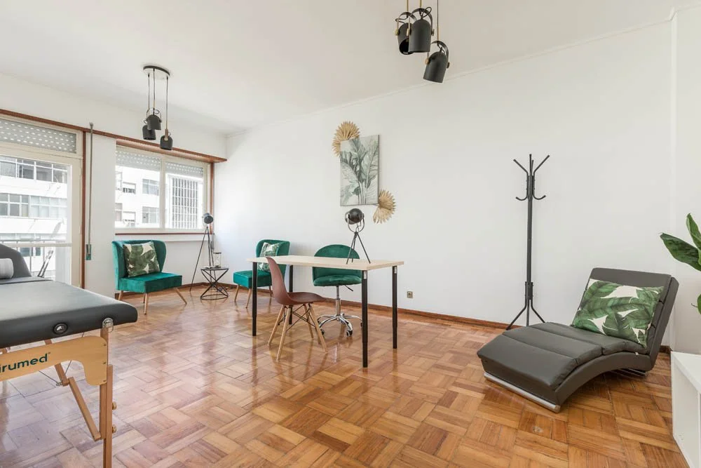 Sala de terapia com poltrona de tratamento, cadeira, mesa, sofá em estilo de chaise, decoração com plantas e quadros na parede, luminárias modernas, piso de madeira.