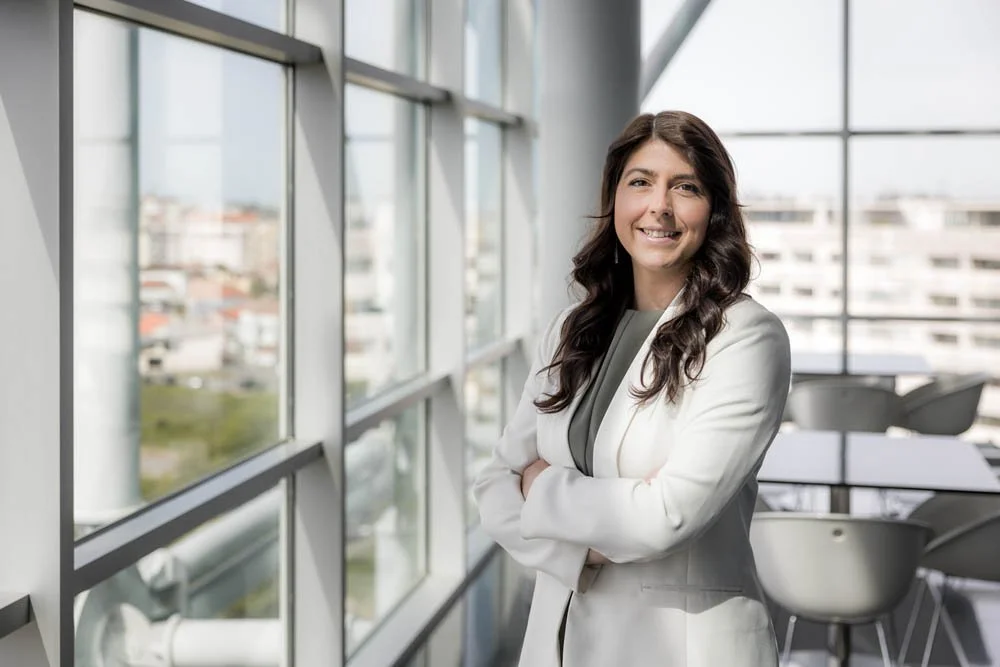 Mulher sorridente vestida com blazer branco, de braços cruzados, no interior de um prédio com janelas de vidro grande e vista para a cidade ao fundo.