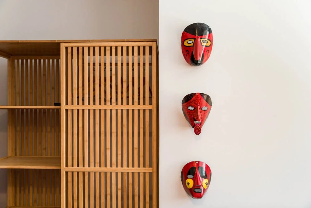 Três máscaras ornamentais penduradas na parede branca ao lado de uma estante de madeira com acabamento liso.