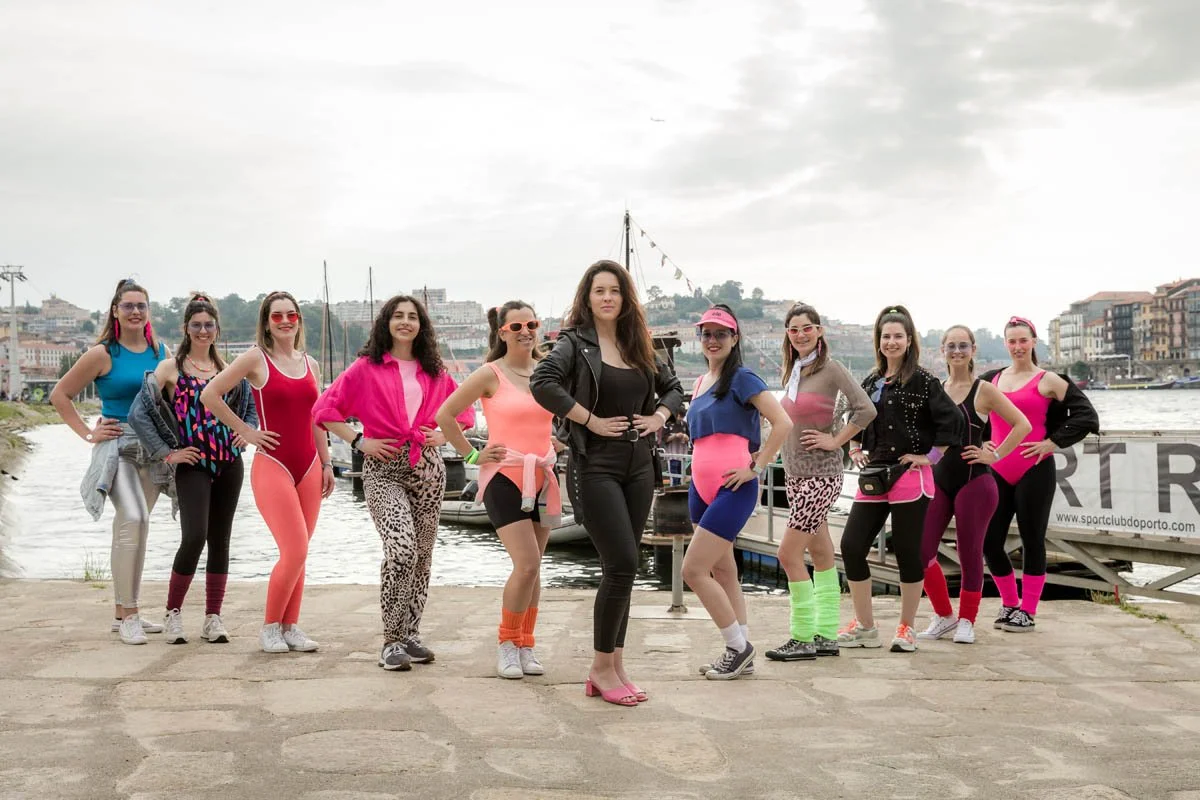 Grupo de mulheres posando na orla do rio, vestidas com roupas de ginástica coloridas e alguns com chapéus e acessórios esportivos, com barcos ao fundo e edifícios ao longo do rio.