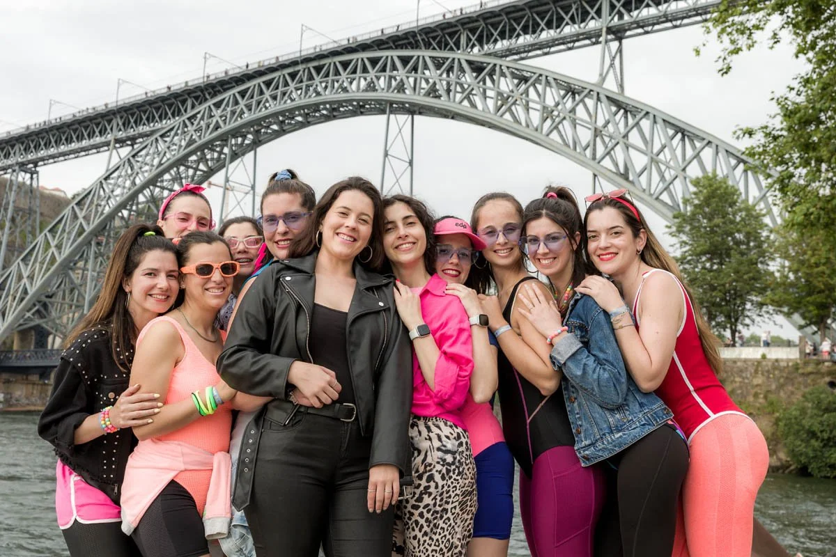 Grupo de mulheres sorrindo posando juntas na rua, com uma ponte de ferro ao fundo, durante o dia nublado.