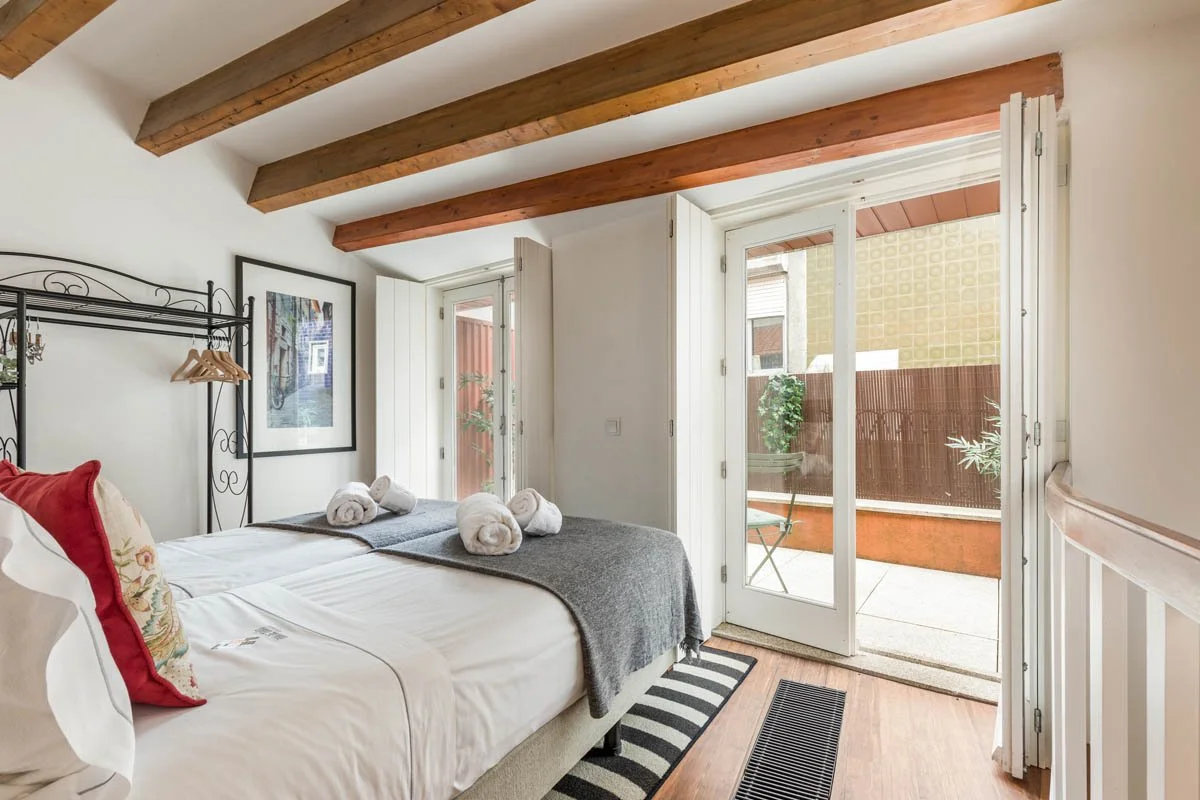 Quarto aconchegante com cama de casal, travesseiros decorativos vermelhos e encosto de cama com estantes de ferro. Decoração com quadro na parede e rolos de toalhas sobre a cama. O quarto tem teto com vigas de madeira expostas e porta de vidro que le