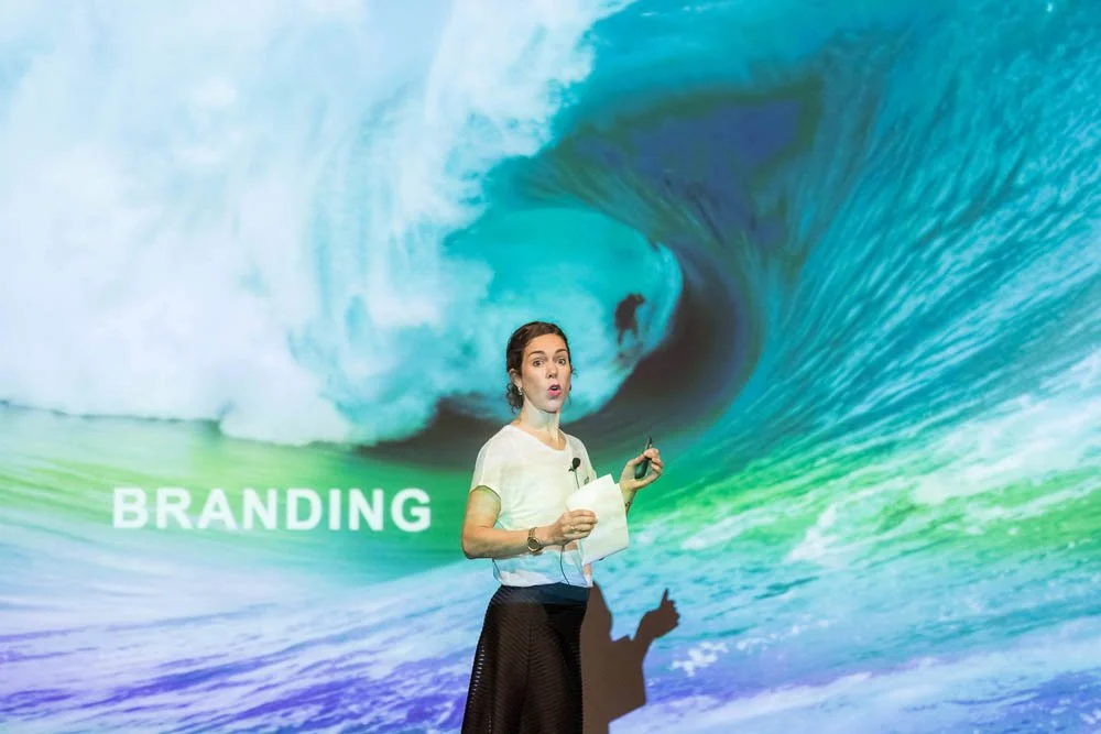 Mulher apresentando em frente a uma produção com a palavra "BRANDING" e uma imagem de uma onda gigante.