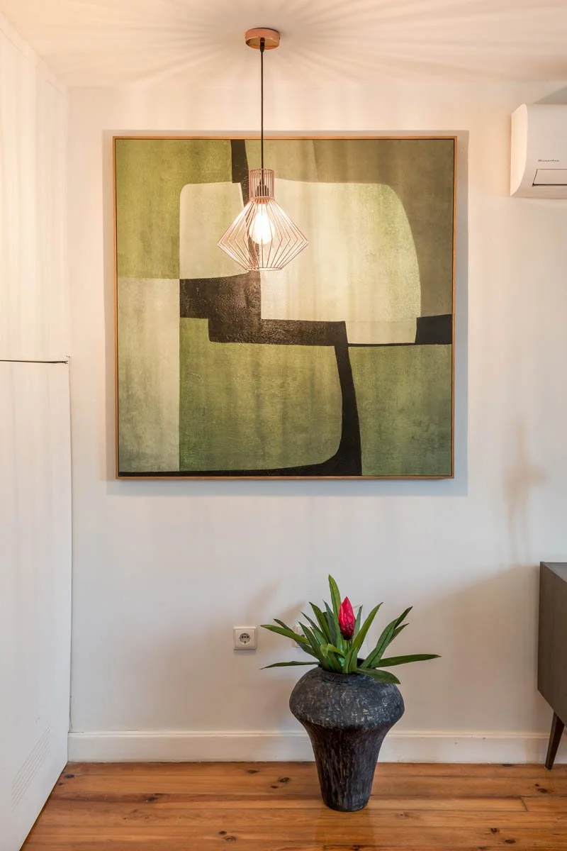 Quadro abstrato em tons de verde na parede, luz pendente acima, vaso com planta e flor na composição da sala.