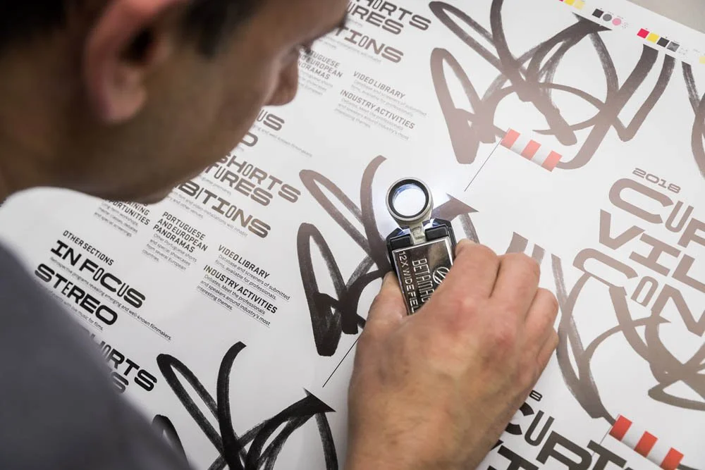 Pessoa usando um espelho para assinar uma folha com várias marcas e anotações, como 'Shorts e Furture' e 'Infectious Stereo', em uma parede ou mesa.