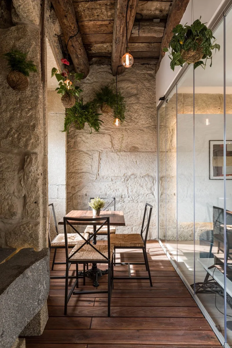 Uma pequena área de descanso com mesa de madeira e quatro cadeiras de metal com assentos de fibra natural, decorada com plantas suspensas e uma planta em um vaso na mesa, paredes de pedra e teto de madeira, separado por uma parede de vidro.