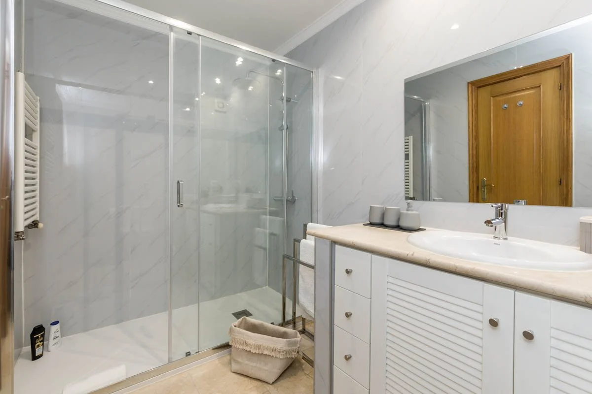 Banheiro com área de banho de box de vidro, pia com espelho grande e armário branco, parede de azulejos brancos e porta de madeira.