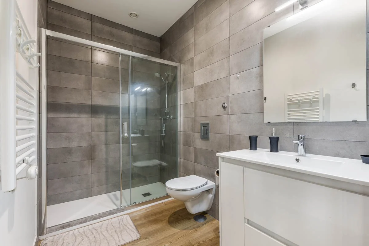 Banheiro com paredes revestidas de azulejos cinzas, box de vidro com chuveiro, vaso sanitário e pia branca com espelho acima. Piso de madeira e toalha branca ao lado do box.