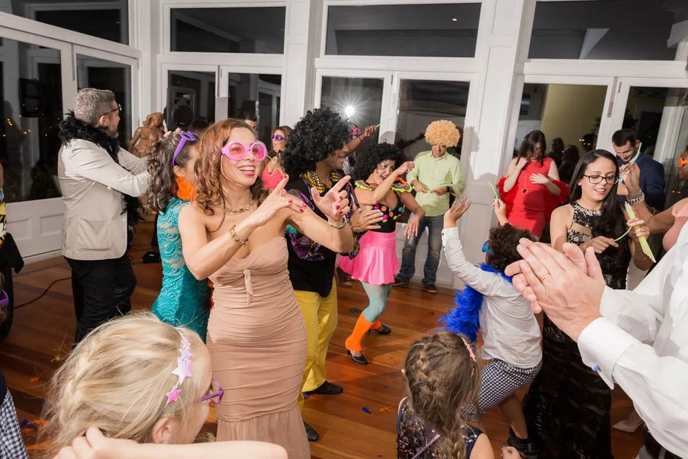 Pessoas celebrando em uma festa com fantasias coloridas e decoração vibrante, dançando e se divertindo.