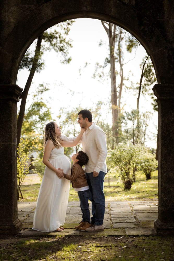 Família feliz na frente de uma estrutura de pedra sob uma árvore em um parque, com mãe grávida, pai, criança e um bebê na barriga da mãe