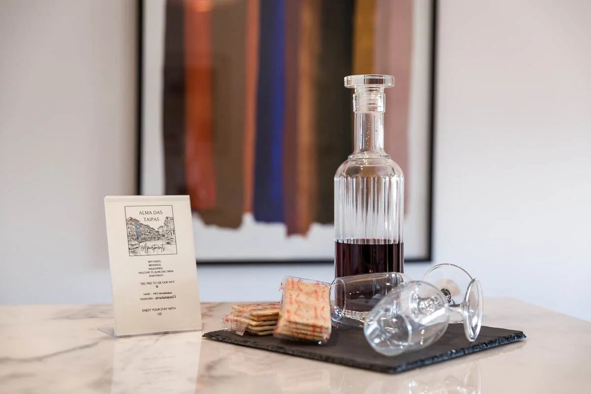 Garrrafa de vidro com vinho tinto, dois copos de vidro deitados em uma bandeja, um pequeno cardápio e pacotes de biscoitos em uma superfície de mármore, com uma pintura de roupas ao fundo.