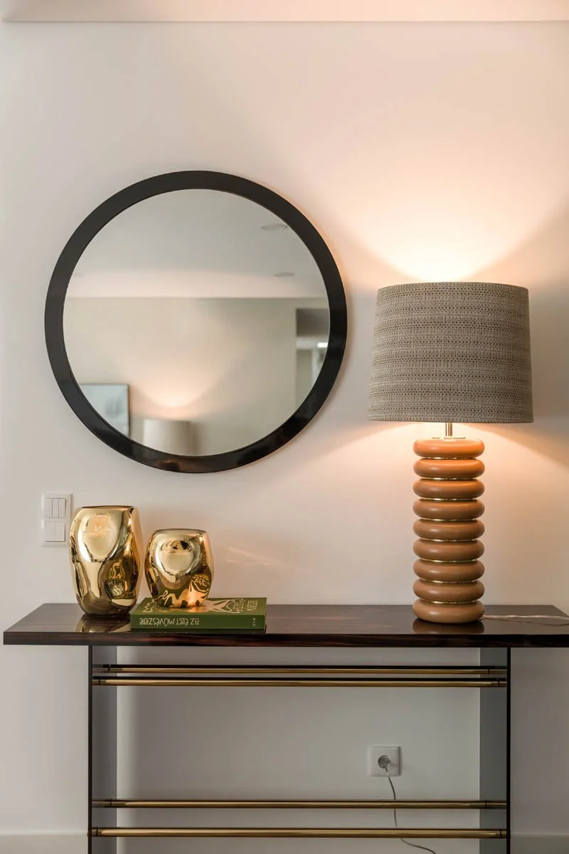 Console com duas esculturas douradas, livro verde, abajur com base decorativa de anéis e espelho redondo na parede.