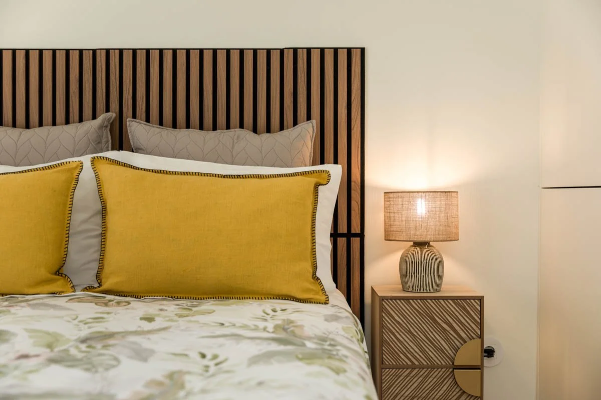 Cama com cabeceira de madeira e almofadas amarelas e bege, ao lado de um abajur com base de cerâmica listrada e luminária acesa, em quarto decorado com cores neutras.