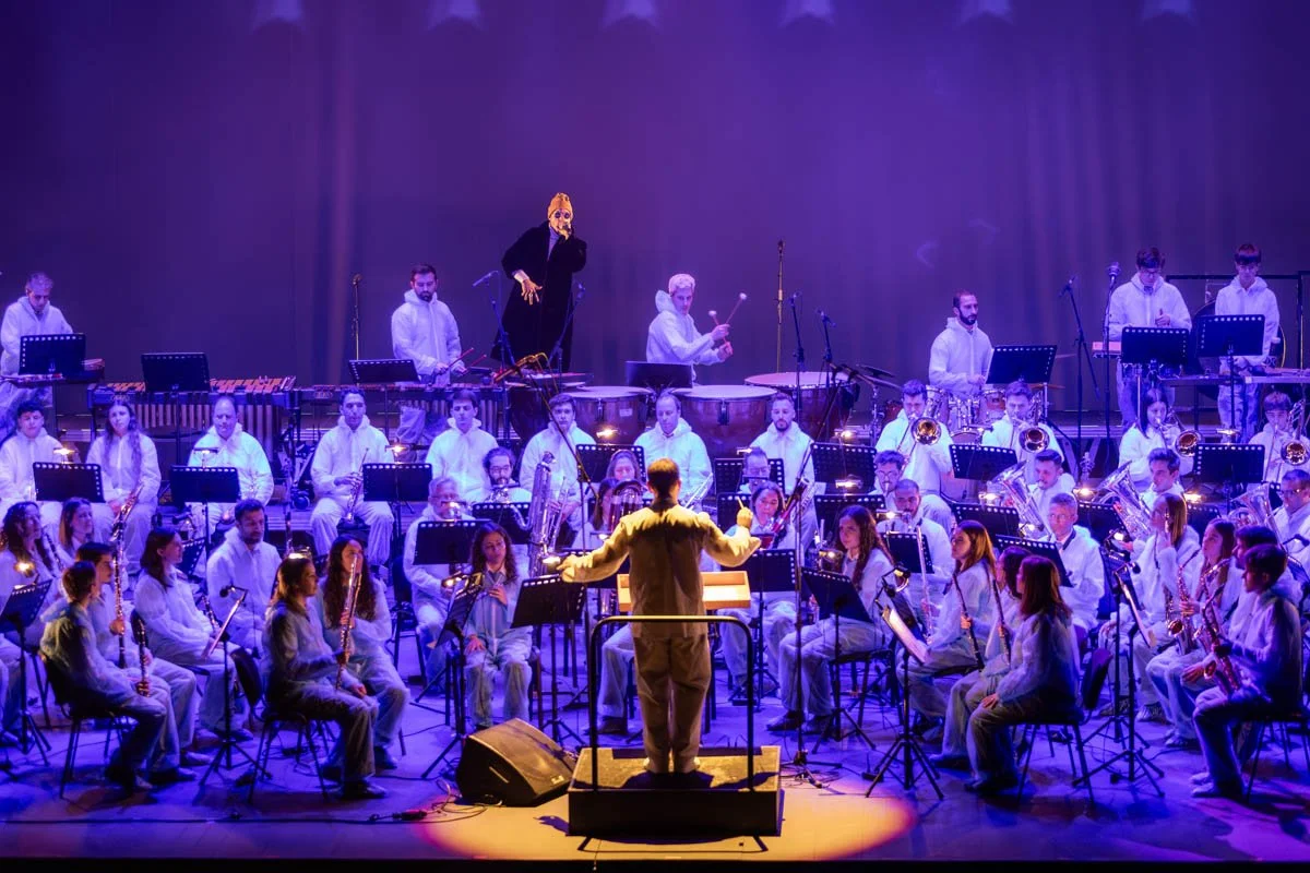 Orquestra de músicos tocando em um palco com iluminação azul e roxa, com maestro ao centro e cantora em destaque ao fundo.