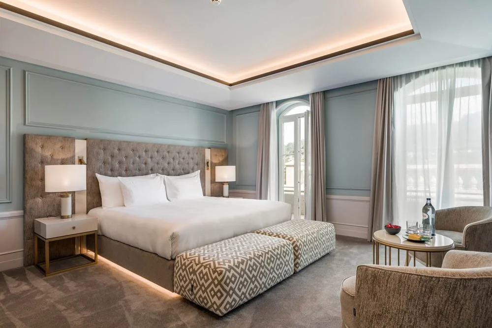Quarto de hotel com cama de casal, cabeceira acolchoada, duas mesas de noite com abajures, poltronas, mesa de centro com água e comida, cortinas na janela e luz de LED no teto.