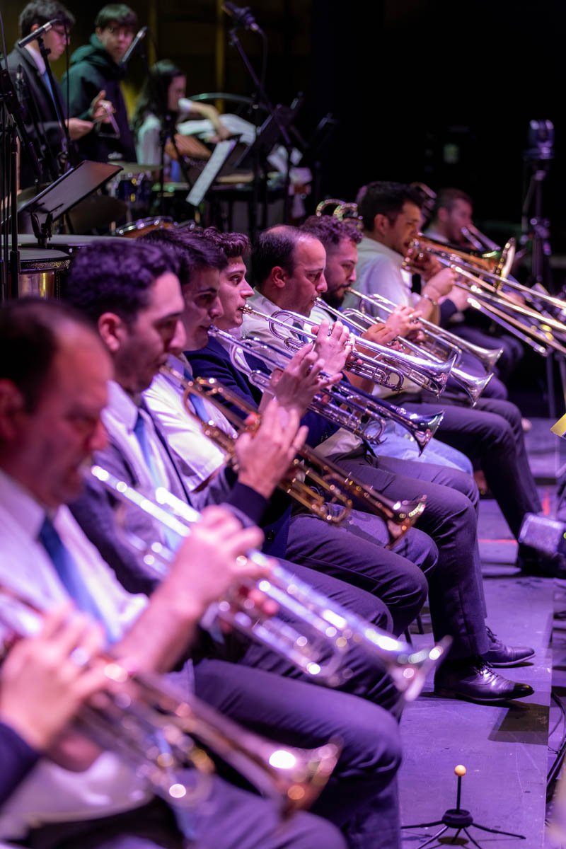 Conjunto de músicos tocando trompetes em uma apresentação ao vivo, com foco na fileira de trompetistas.