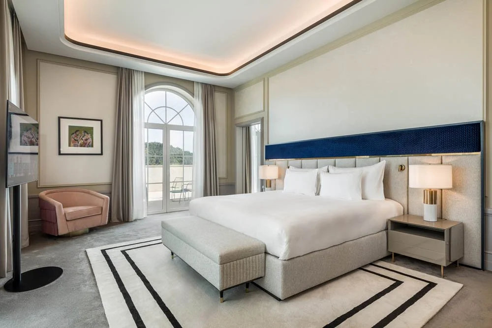 Quarto de hotel com cama de casal, cabeceira azul, dois abajures ao lado, mesa de cabeceira, poltrona rosa, televisão na parede, janelas com cortinas e porta de vidro para varanda com vista para o exterior.