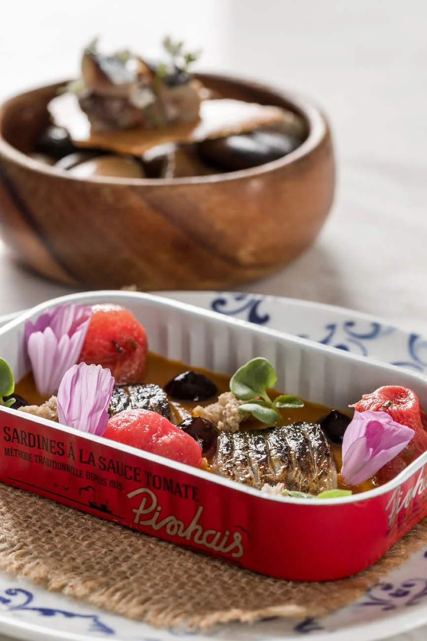 Lata de sardinhas ao molho de tomate decorada com flores e folhas, na frente uma tigela com pratos de comida e uma tigela de madeira contendo ingredientes diversos.