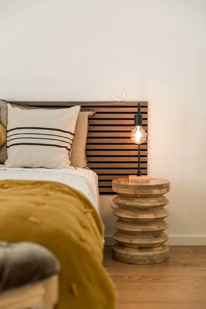 Quarto com cabeceira de cama de madeira com painéis horizontais, travesseiros e uma manta amarela. Ao lado, mesa de cabeceira de madeira com design em camadas, luminária com lâmpada exposta.