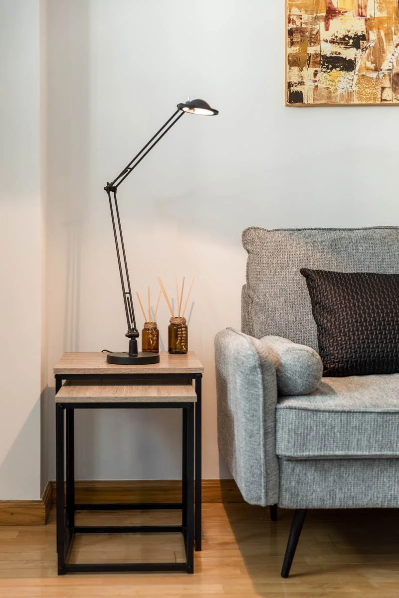 Sala de estar com sofá cinza, mesa lateral de madeira com estrutura preta, luminária de mesa ajustável, duas garrafas com sticks aromáticos e uma pintura abstrata na parede