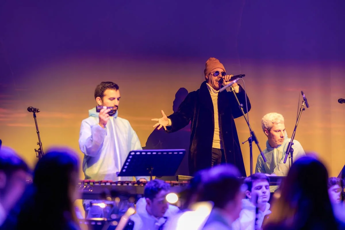 Performers no palco com instrumentos musicais durante um show, com público assistindo na frente.